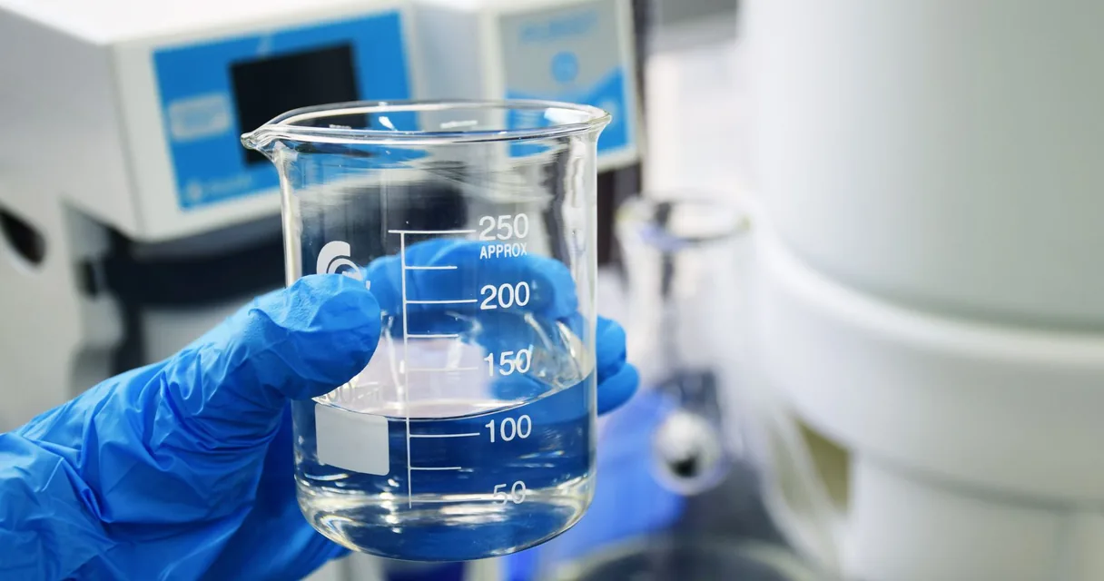 Béquer com água deionizada segurado por mão com luva azul em laboratório
